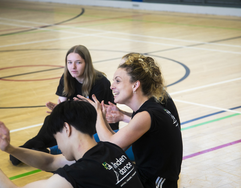 white female teacher with teenage student sitting on a sports hall floor with hands gesturing. All wearing black with linden dance logo on the t-shirts. 