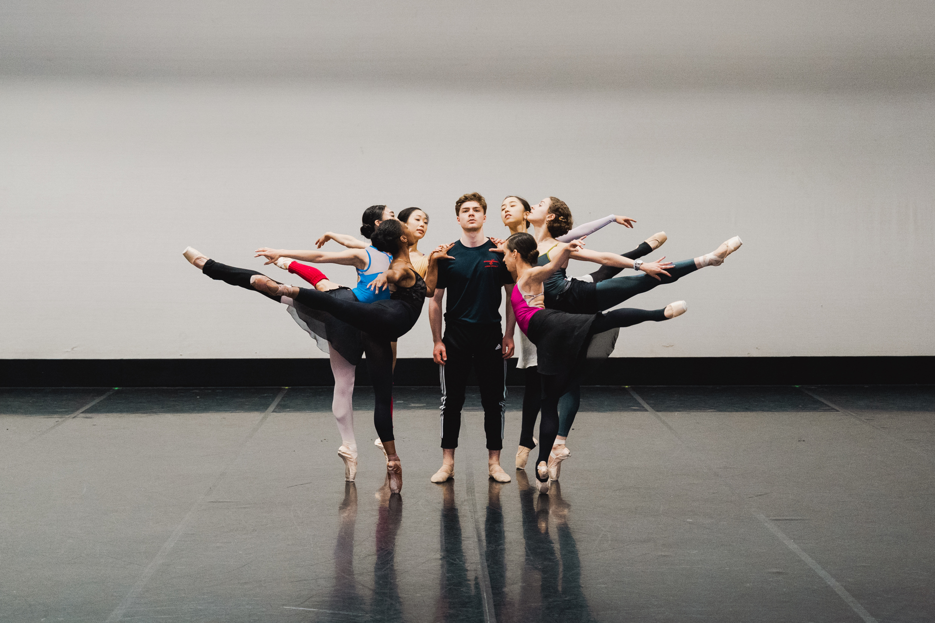 six females on point in an arabesque with leg high behind all holding onto centre male who is standing looking at the camera. In a dance studio 