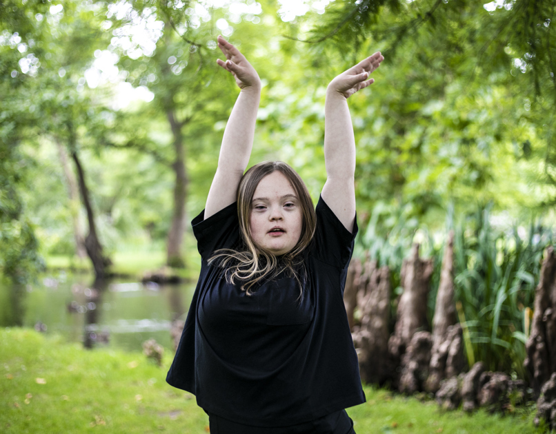 Blonde white female down's syndrome dancer wearing all black. Both hands raised above her head looking right at the camera, with soft focus trees in the background