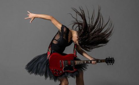 female dancer on point with bended knees, holding a guitar arching over it, with one hand flung behind and long brown hair flicked over the head