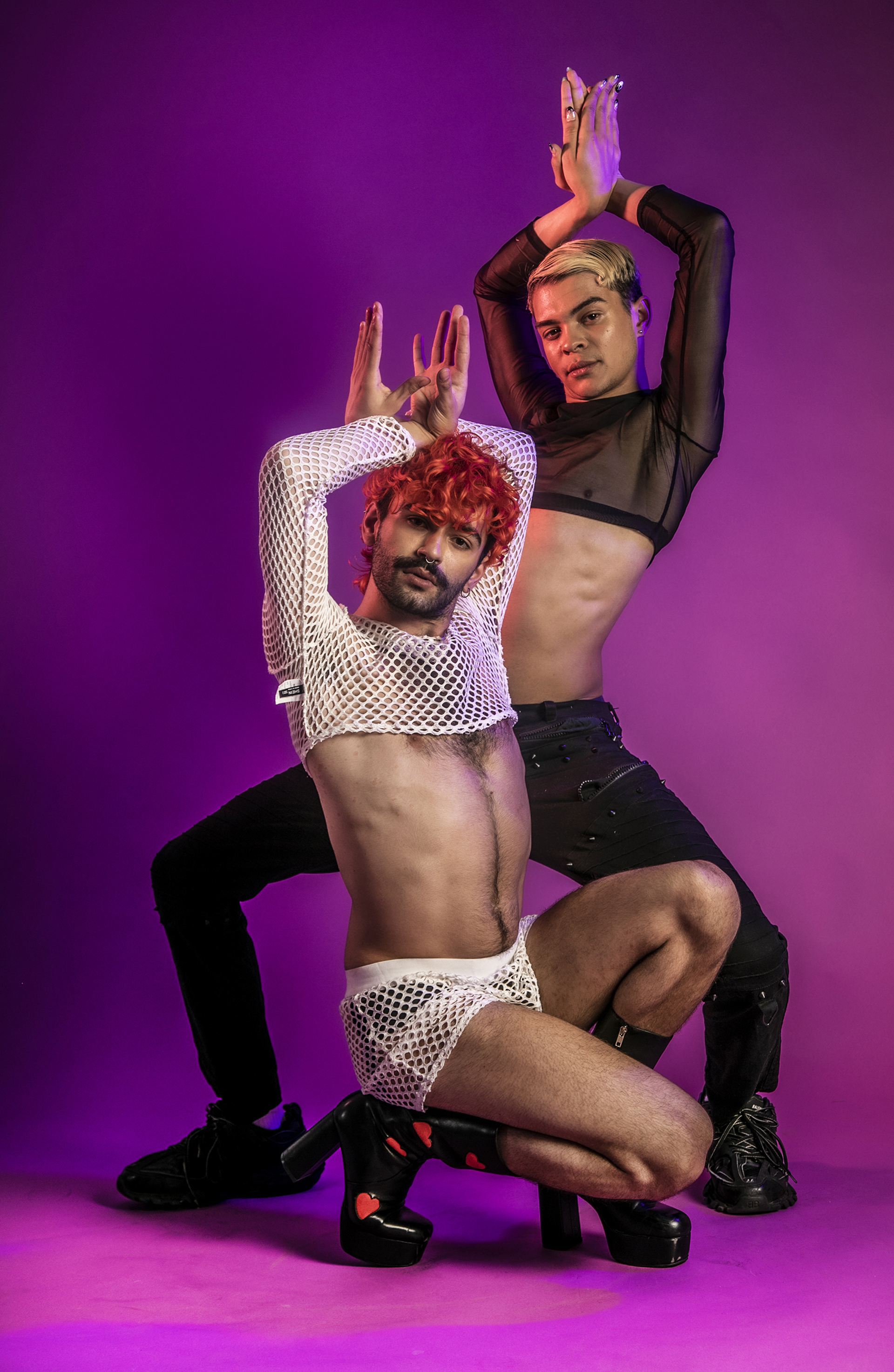 Two global majority LGBTQ+ male voguing dancers, one crounched down on knees with hands met above head and the other in a half squat behind with hands met above head . Wearing white and black net and platform shoes on pink studio background. 