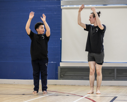 teenage male dance teacher reaching up to the sky with both arms teaching young boy dancer who is mirroring the arms in the air. In sports hall both wearing black PE kit