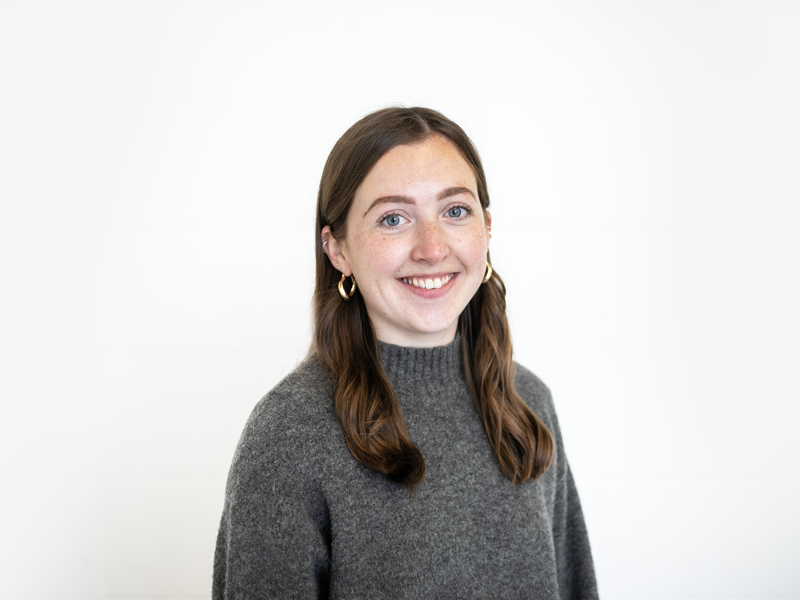 Headshot of Chloe Sprackling white female with brown hair smiling at the camera. Wearing gold hoop earrings and high neck grey jumper