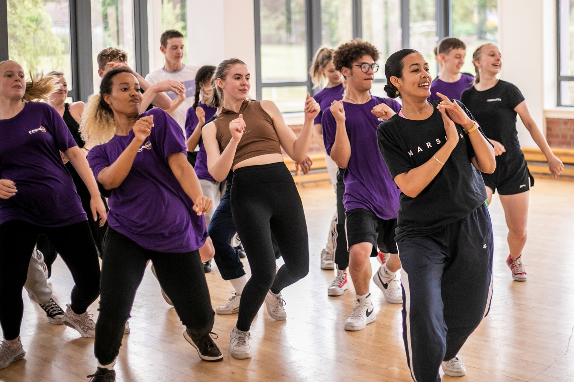 Female global majority dancer with curly hair tied back teaching a hip hop class. Students copying the lean back movement behind all wearing purple t-shirts in brightly lit dance studio