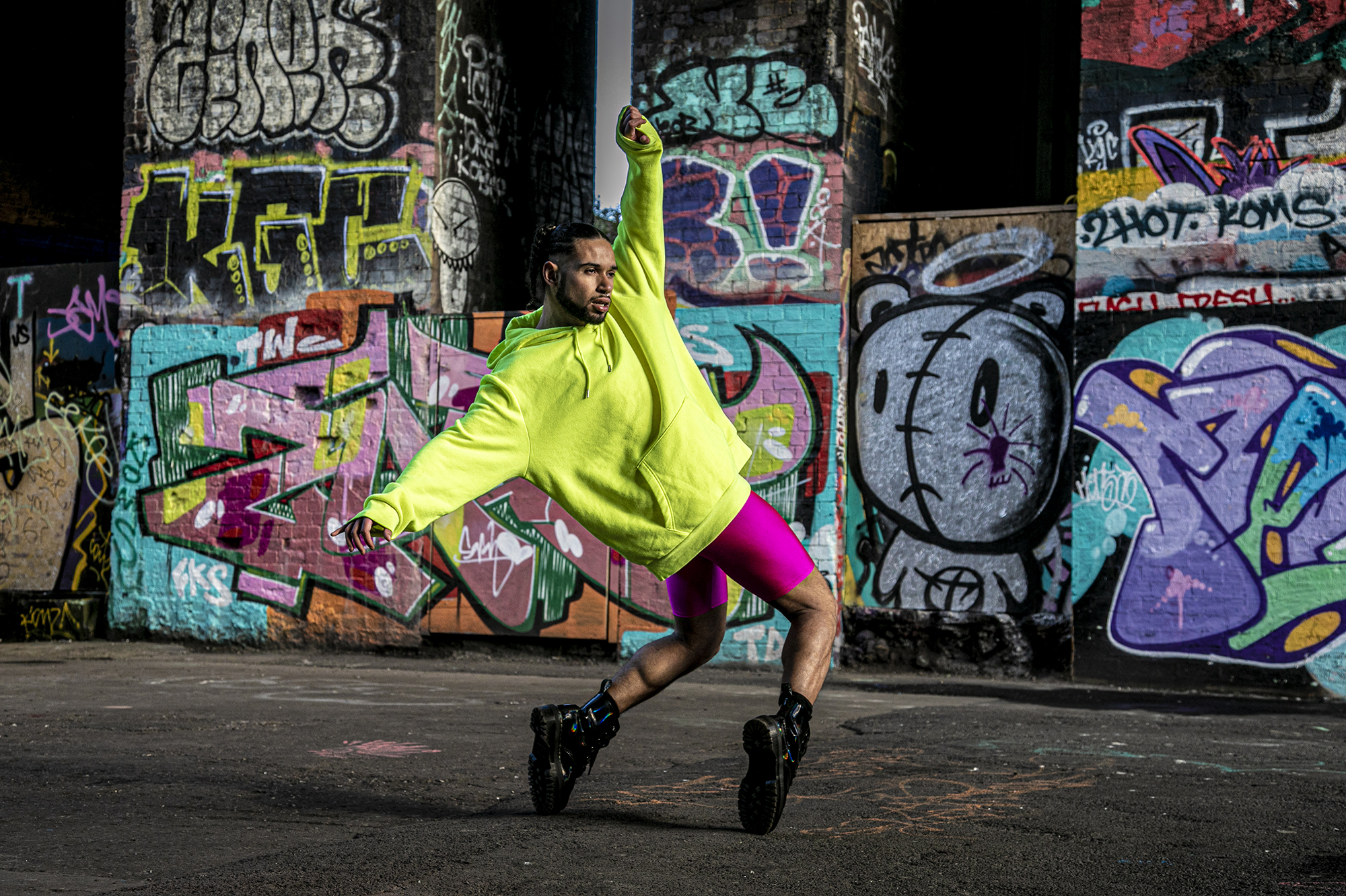 LGBTQ+ global majority male dancer with short dreads wearing neon yellow jumper with pink leotard shorts and black boots. Hands around the face, standing top toes with bent knees leaning back with arms out stretched. In front of colourful graffiti wall.