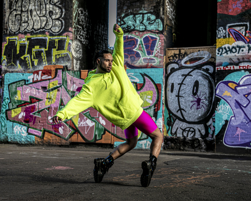 LGBTQ+ global majority male dancer with short dreads wearing neon yellow jumper with pink leotard shorts and black boots. Hands around the face, standing top toes with bent knees leaning back with arms out stretched. In front of colourful graffiti wall.