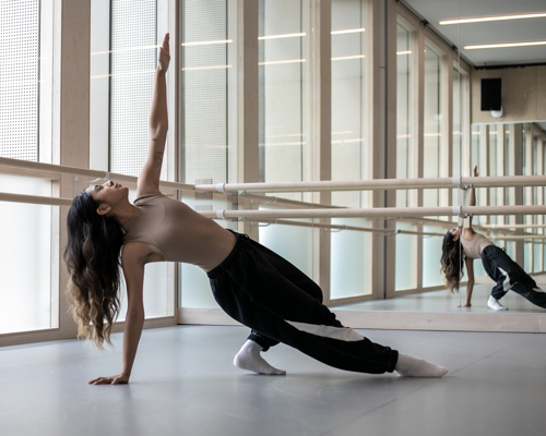 Global majority female dancer with nude coloured crop top with black trousers.  One arm up and the other holding her up from the floor with one extended leg and one bent leg. White mirrored dance studio 