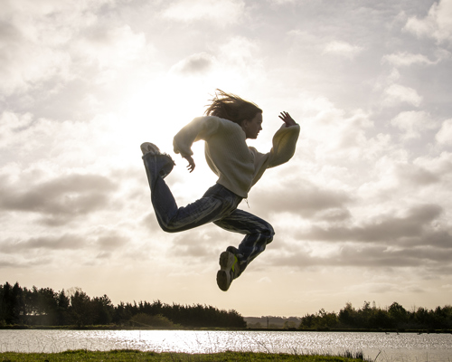 white female dancer jumping in the air in a running shape in front of golden clouded sky wearing casual clothes