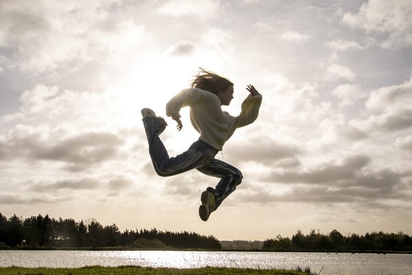 white female dancer jumping in the air in a running shape in front of golden clouded sky wearing casual clothes
