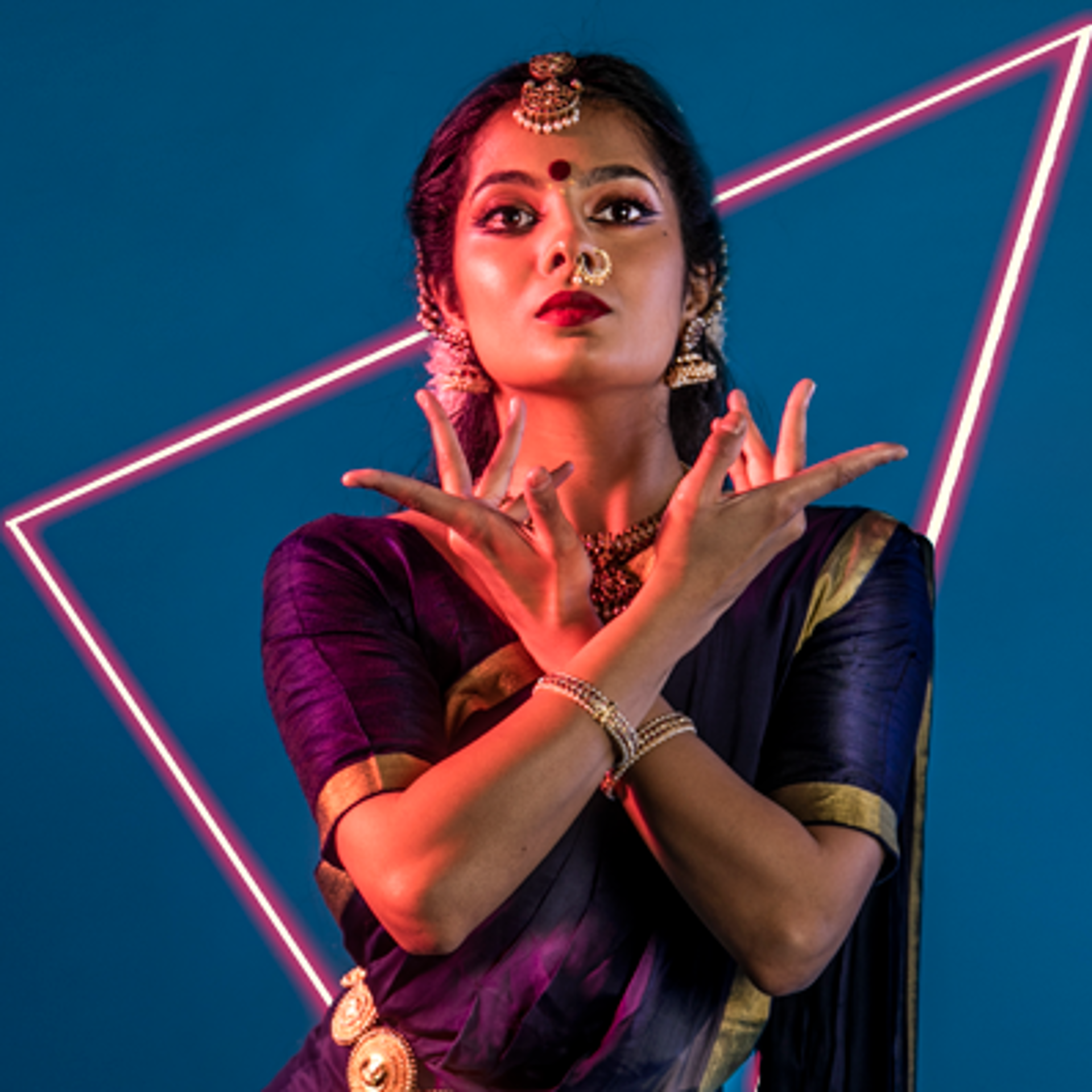 South Asian dancer holding both hands crossed over her chest and under her chin looking to the topof the screen. Wearing traditional purple dress with gold belt bracelets necklace and head jewlery with gold nose ring. Bright purple studio background.