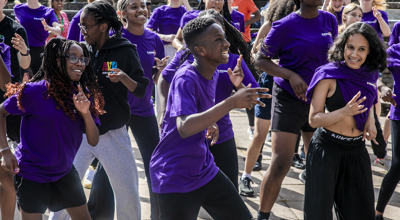 Young people dancing in an outdoor warm up, central dancer is a black male smiling with hands waving infront of him. Wearing a mix of coloured outfits. 