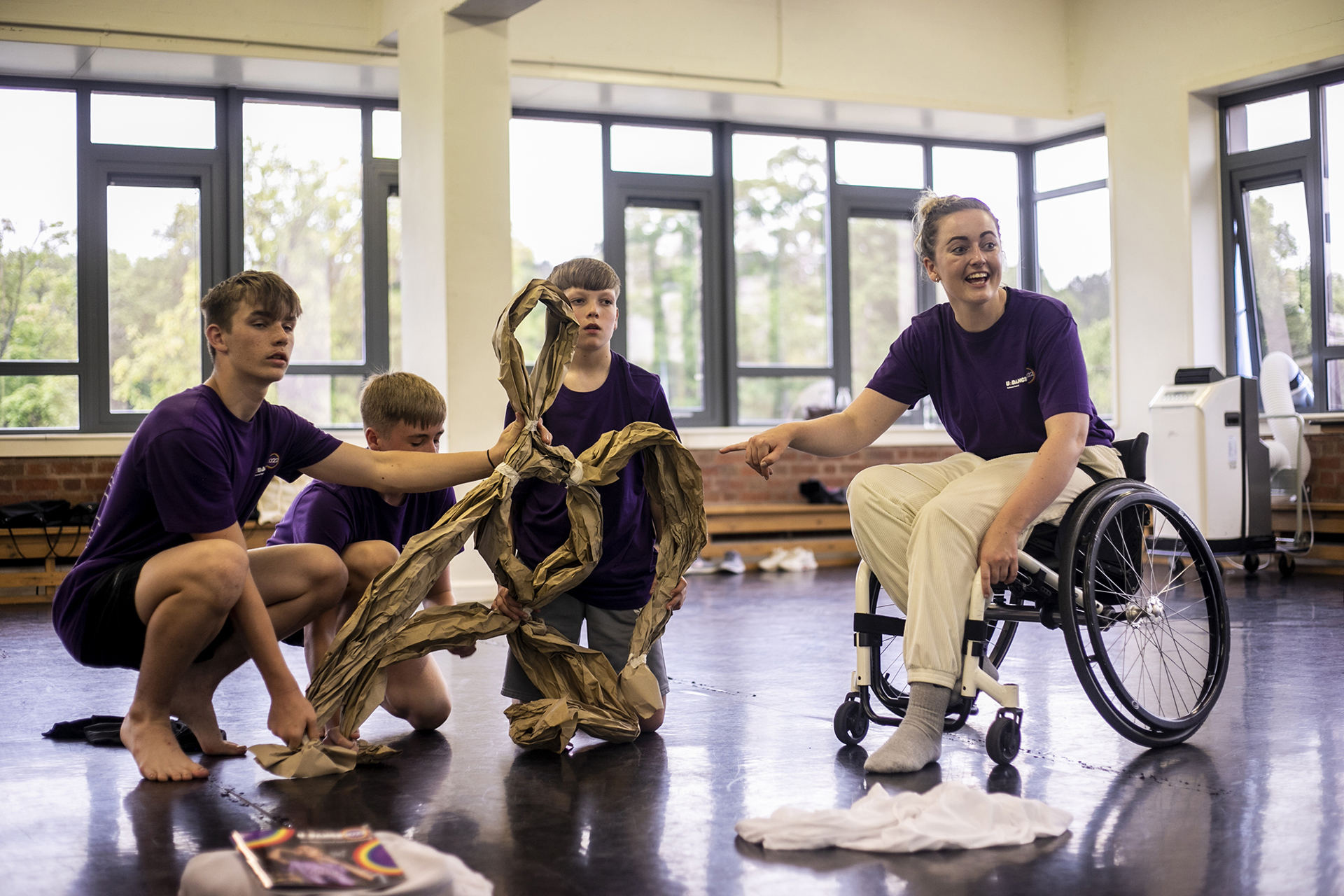 three young white boys holding up a paper puppet with a white female wheel chair dancer holding the puppets arms and smiling, in a dance studio.