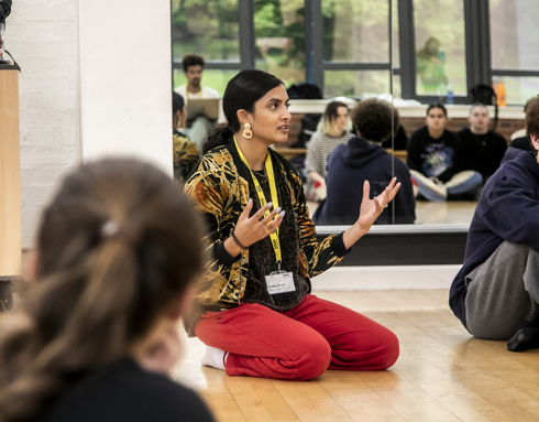 golbal majority dance teacher sitting on her knees speaking to a room full of dance students gesturing her hands, with mirror reflecting the students behind her. Wearing red trousers and colourful jacket. 