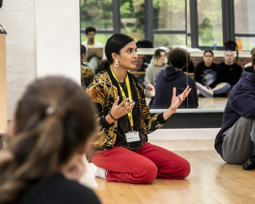 golbal majority dance teacher sitting on her knees speaking to a room full of dance students gesturing her hands, with mirror reflecting the students behind her. Wearing red trousers and colourful jacket. 