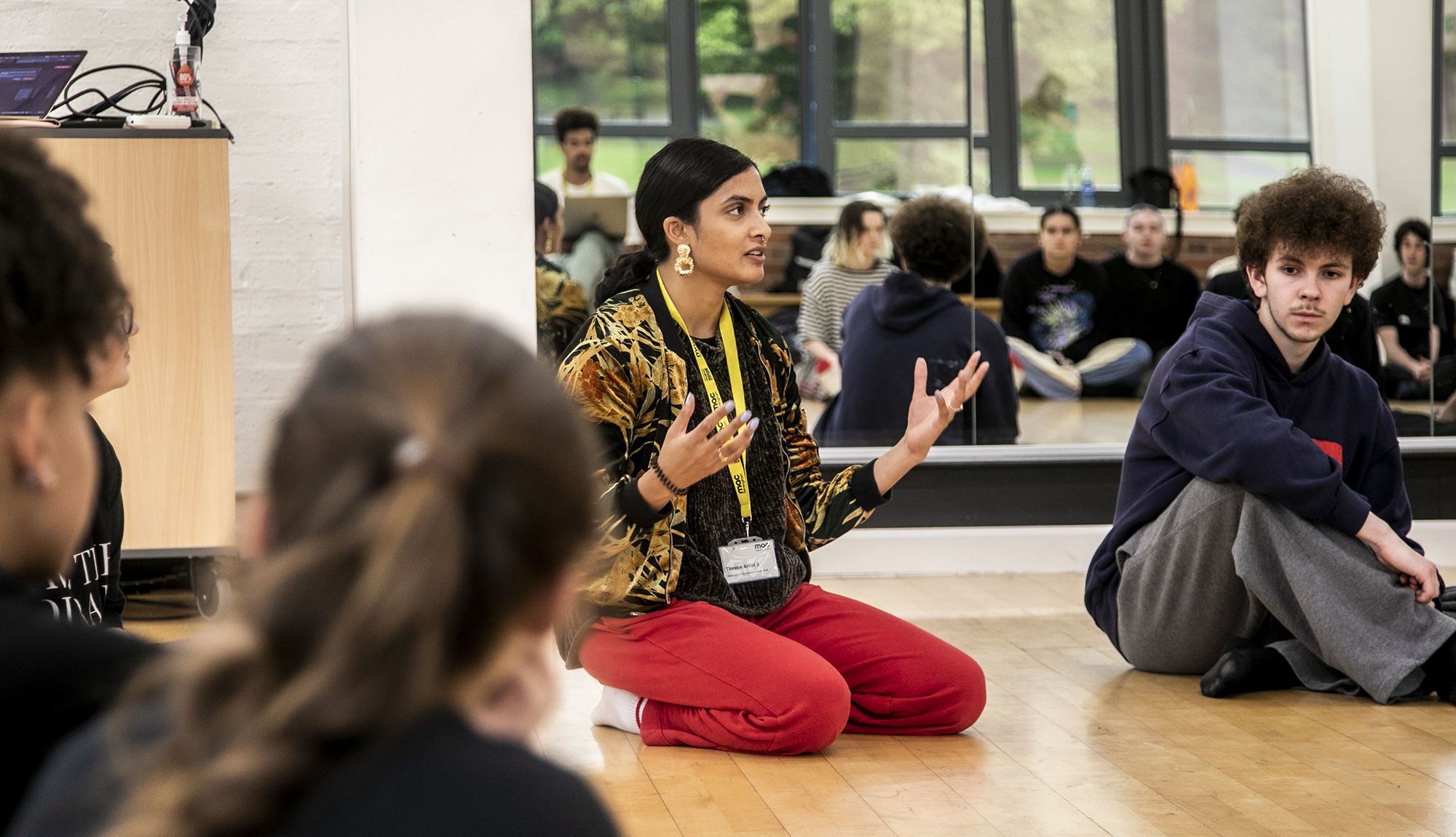 golbal majority dance teacher sitting on her knees speaking to a room full of dance students gesturing her hands, with mirror reflecting the students behind her. Wearing red trousers and colourful jacket.  golbal majority dance teacher sitting on her knees speaking to a room full of dance students gesturing her hands, with mirror reflecting the students behind her. Wearing red trousers and colourful jacket.