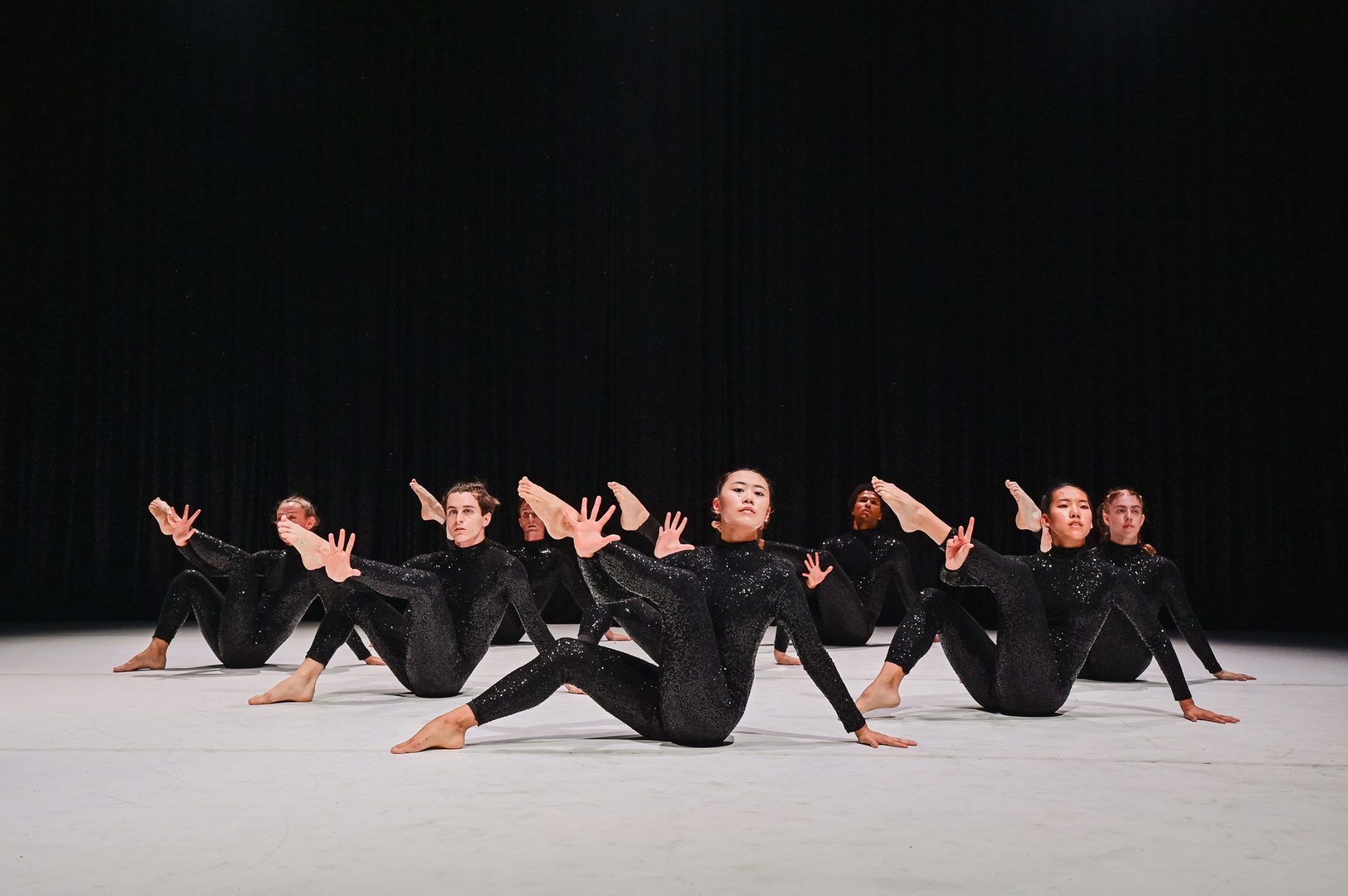 Seven dancers sat on the floor holding a leg up to the left of the image with the other leg bent with one hand on the white floor . Wearing black sequin bodysuits, with a black background 