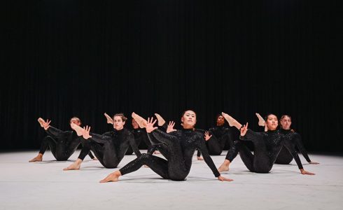 Seven dancers sat on the floor holding a leg up to the left of the image with the other leg bent with one hand on the white floor . Wearing black sequin bodysuits, with a black background 