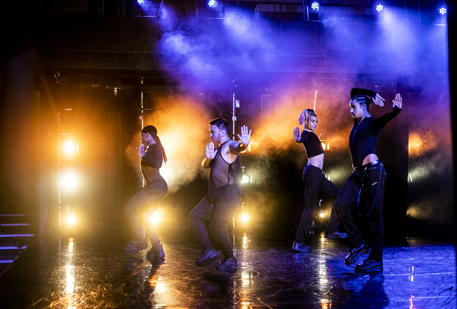 Side of the stage looking into orange stage lights with a line of four vogueing LGBTQ+ dancers with to the side of them, wearing black with bright purple orange and blue lighting behind them