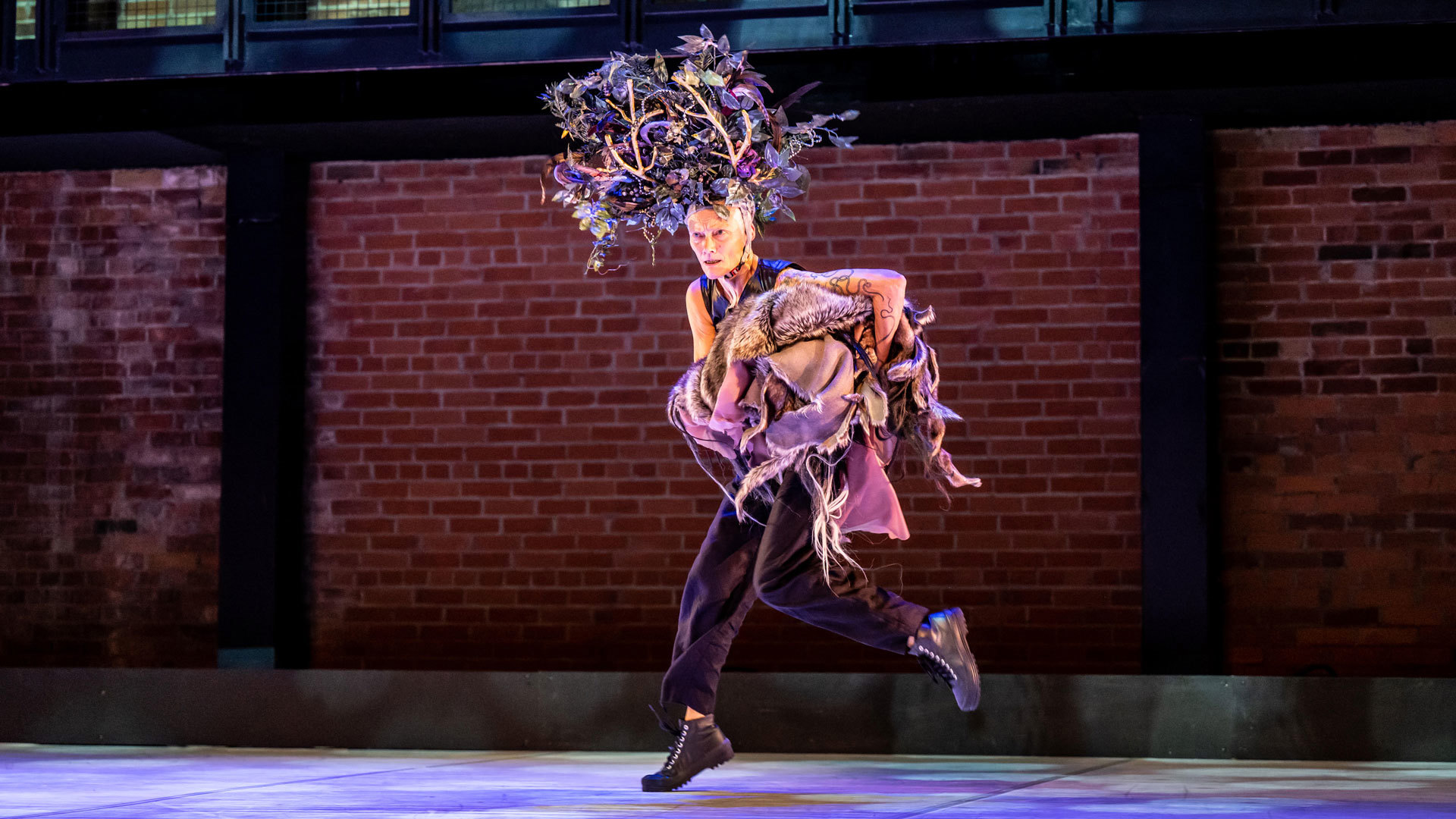 white female dancer running across the stage with large tree head piece carrying lots of clothes with brick background
