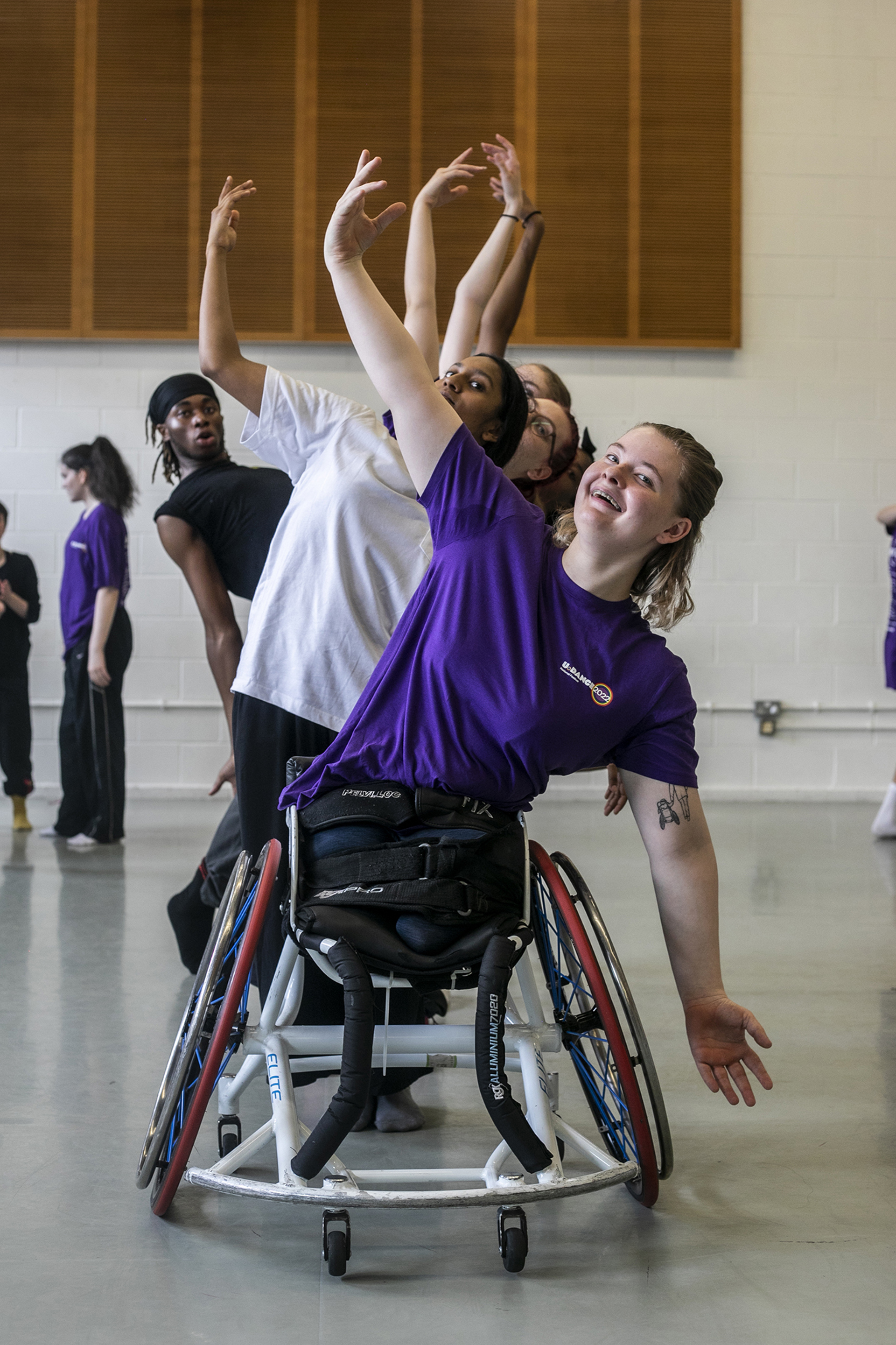 White female dancer in a wheel chair lifting one arm above her head with a mix of male and female, different raced dancers standing behind doing the same lifting arm pose, in a dance studio