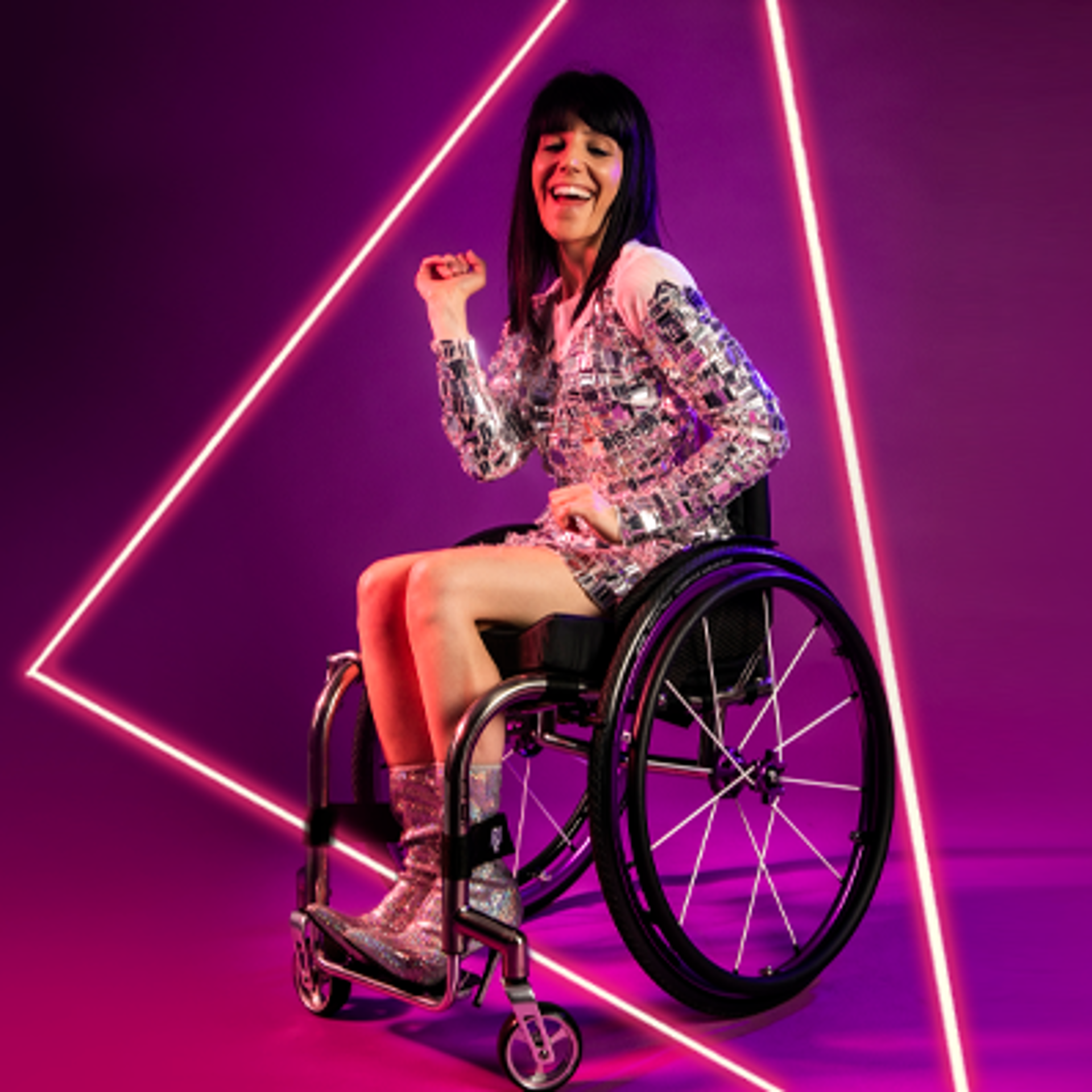 White female wheelchair dancer with black hair. With one arm up and one down with wheel chair side on. Wearing silver sparkle dress and silver high heeled shoes in front of hot pink studio background.