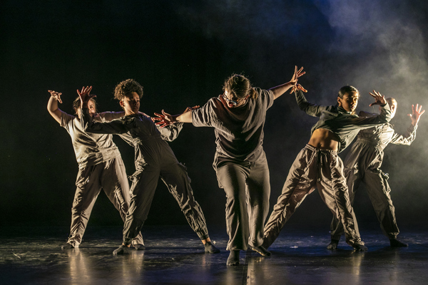 five female dancers one in the center with arms puhing out the other four leaning back from the push with arm out to the side. One a dark stage with smoke over back light on the right hand side. All wearing dark neutral tones