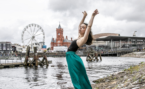 dancer with arched back and arms above head infront of Cardiff bay skyline. Young white female wearing black leotard and green silk trousers