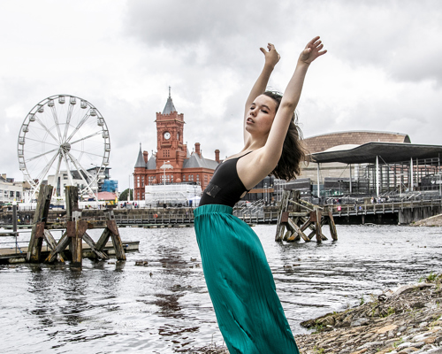 dancer with arched back and arms above head infront of Cardiff bay skyline. Young white female wearing black leotard and green silk trousers