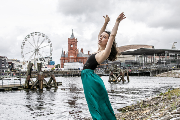 dancer with arched back and arms above head infront of Cardiff bay skyline. Young white female wearing black leotard and green silk trousers