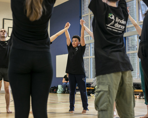 Dance circle with young people holding hands with the joint hands in the air. All wearing black PE kit in a sports hall.