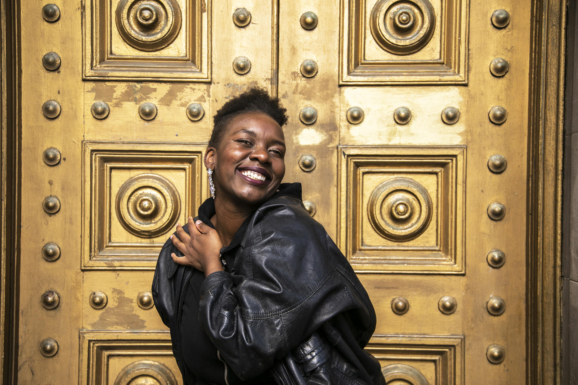 global majority female dancer holding her heart smiling at camera wearing black leather jacket with short afro hair in front of gold door. 