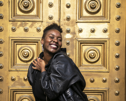 global majority female dancer holding her heart smiling at camera wearing black leather jacket with short afro hair in front of gold door. 