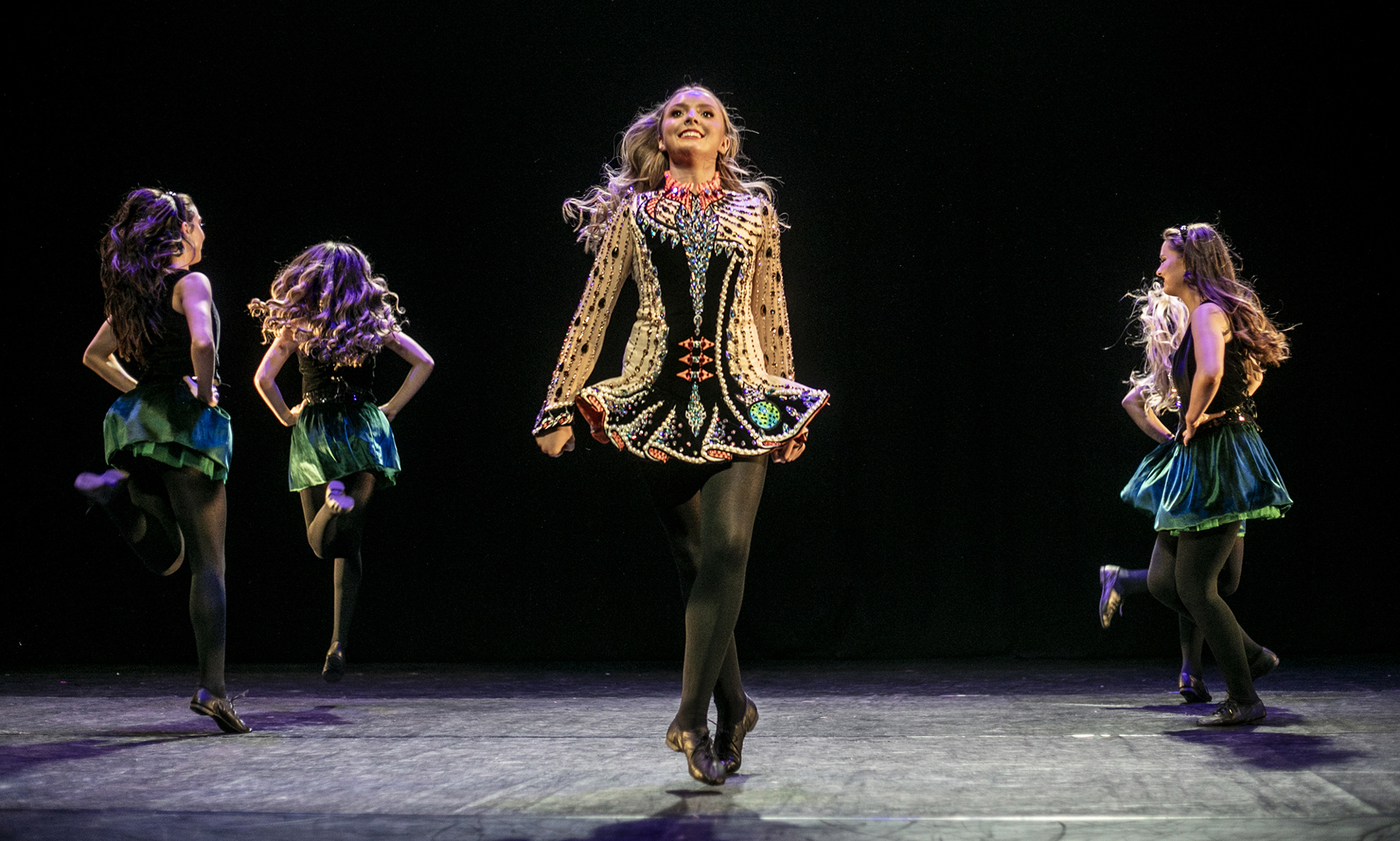 White female Irish dancers circleing one female dancer with arms and legs straight with feet crossed wearing gold traditional Irish dress. Background dancers wearing black and green dresses on a dark stage 