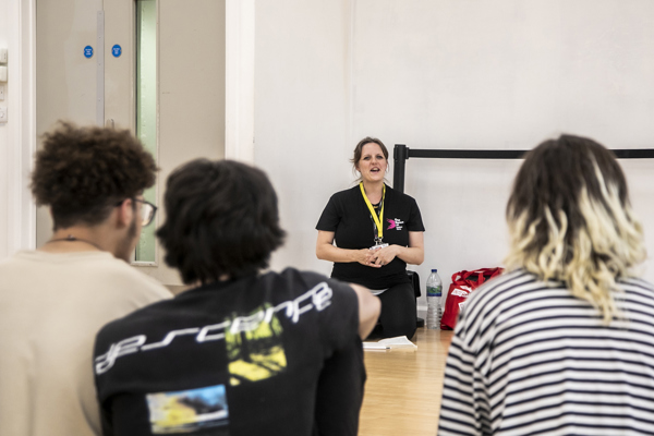 One Dance UK staff member sitting on knees talking to three students sitting in front of the camera in a dance studio. 