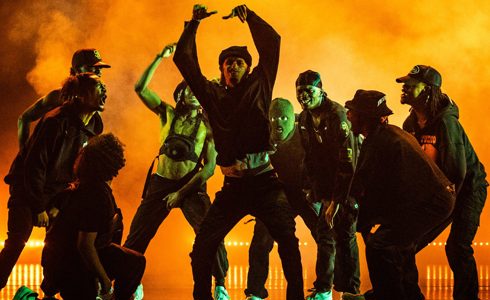 Nine male dancers huddled around eachother shouting and cheering at central dancer who has his arms in the air. All wearing dark clothes on a dark stage with orange light over smoke