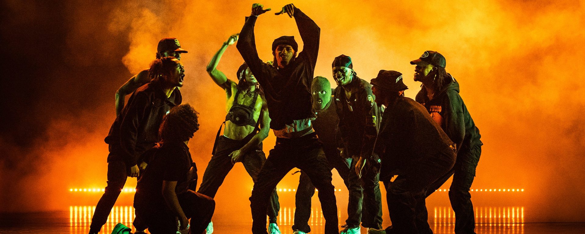 Nine male dancers huddled around eachother shouting and cheering at central dancer who has his arms in the air. All wearing dark clothes on a dark stage with orange light over smoke